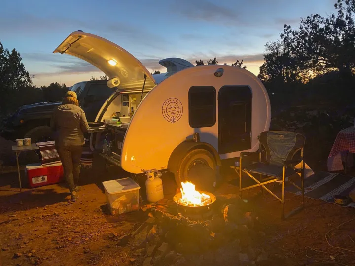 Enjoying an RV rental adventure! A teardrop camper with an open galley, a crackling campfire, and a person setting up camp at dusk.. Bean Bean Stock 2021