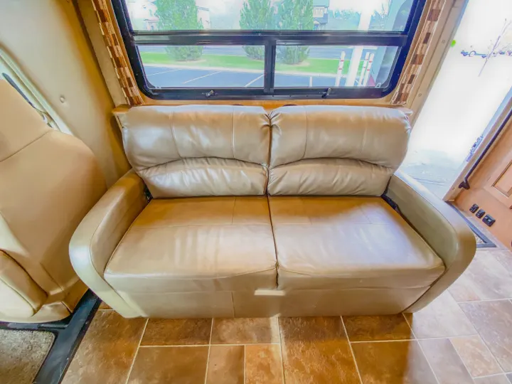 Relax in this RV rental's spacious living area, featuring a tan leather sofa and large window offering scenic views.. Thor Chateau 2016