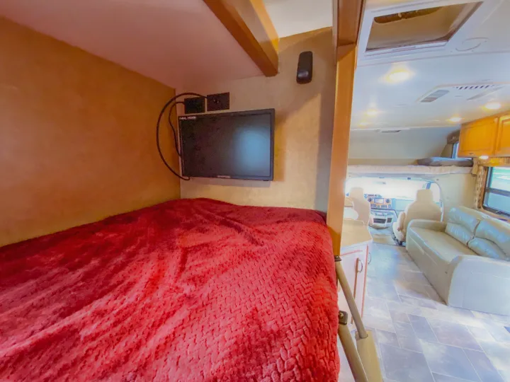 RV rental interior: A bunk bed with a red blanket and mounted TV is in the foreground, with the spacious living area visible behind.. Thor Chateau 2016