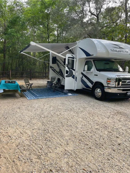 White Magnitude RV rental at a gravel campsite. Awning extended, with outdoor chairs, a table, and a blue rug.. Thor Magnitude GA22 2022