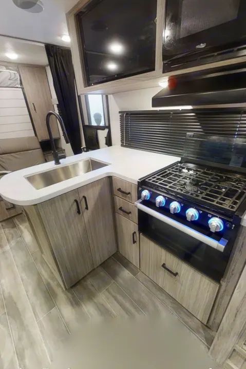 Modern RV rental kitchen with U-shaped counter, sink, stove, oven, microwave, and TV, all within light wood-grain cabinets.. Keystone RV Keystone Passport 2021