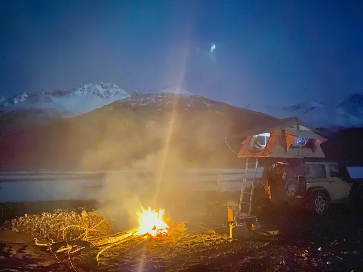 A warm bonfire lights a dark night campsite, revealing an RV rental with a cozy rooftop tent and snowy mountains.. Jeep Rubicon 2014