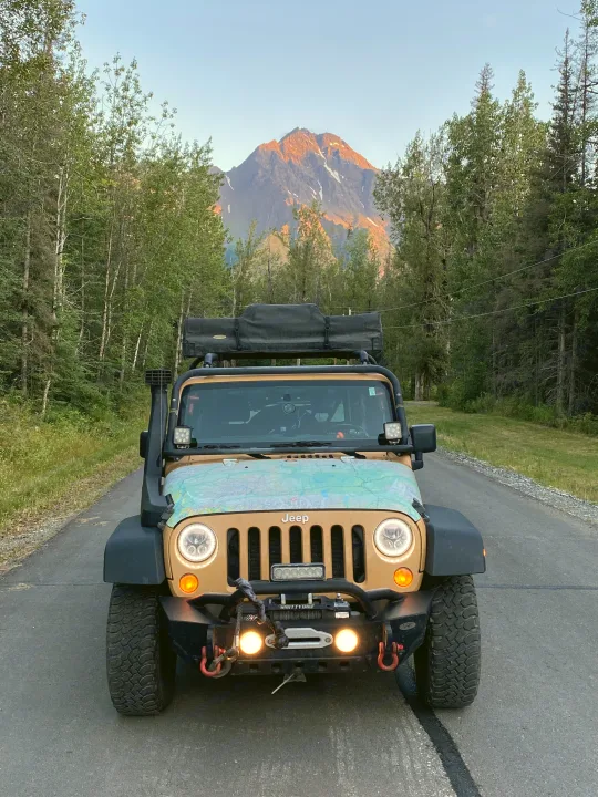 A tan, modified Jeep Wrangler, great for an RV rental adventure, on a road with a majestic, sunlit mountain and dense forest.. Jeep Rubicon 2014
