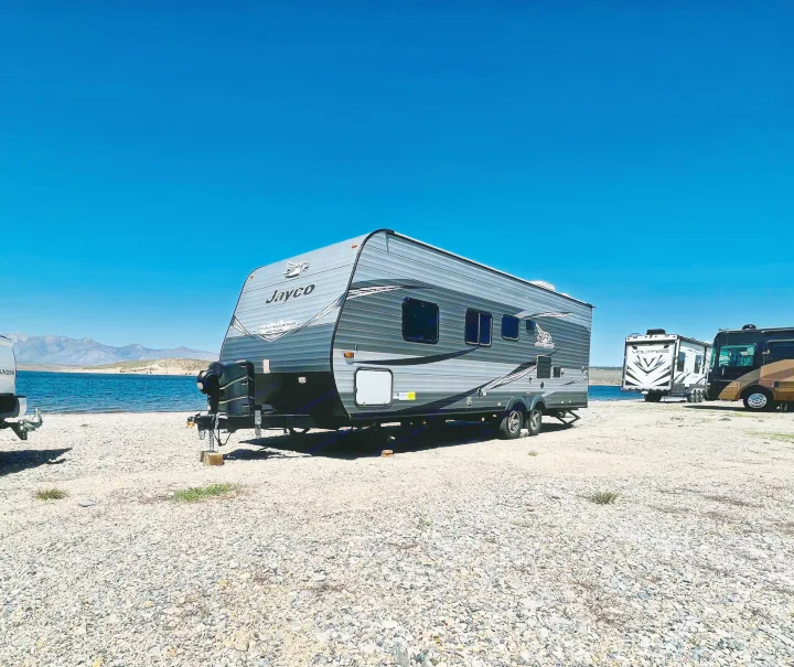A grey Jayco travel trailer (RV rental option) parked on a rocky lake shore with mountains under a clear blue sky.. Jayco Jay Flight SLX Rocky Mountain Edition 2021