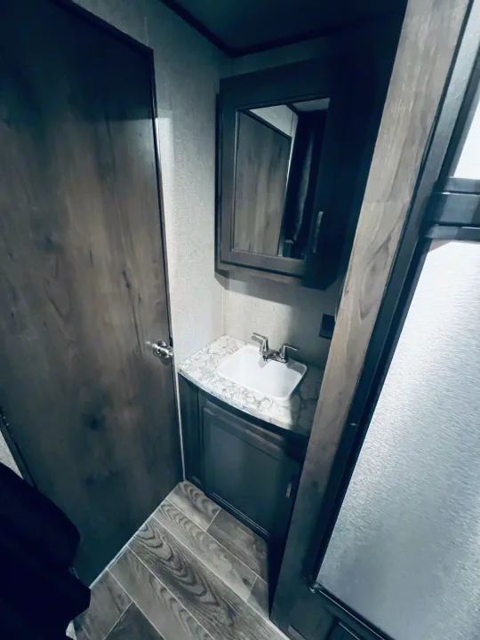 Compact RV rental bathroom with wood-look floors, a sink, vanity, and mirrored cabinet. Includes a shower entry on the right.. Jayco Jay Flight SLX Rocky Mountain Edition 2021