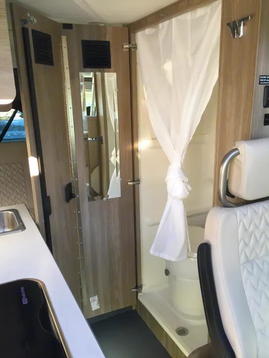 RV rental interior showing a compact shower with curtain, white quilted seating, and a fold-down counter with mirrors.. Winnebago Solis 2022