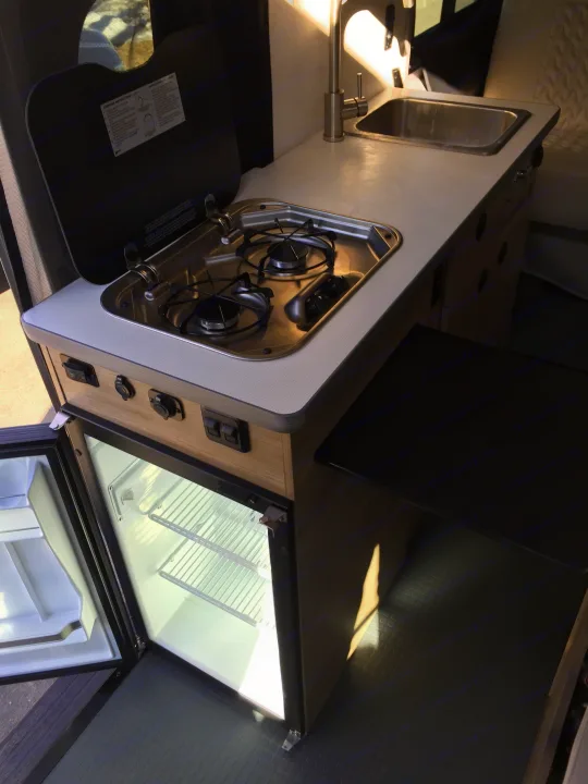 Inside an RV rental: a compact kitchen with a sink, two-burner stove, and an open refrigerator.. Winnebago Solis 2022