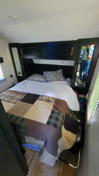 Cozy bedroom inside an RV rental, with a plaid quilt, dark headboard, and mirrored cabinets. A person is visible in the reflection.. Forest River Cherokee 2022