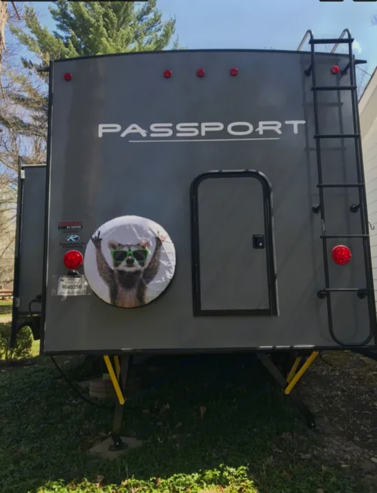 Rear of a grey Passport RV, ready for your next RV rental trip! Features a fun raccoon tire cover, ladder, & stabilizer jacks.. Keystone RV Keystone Passport 2021