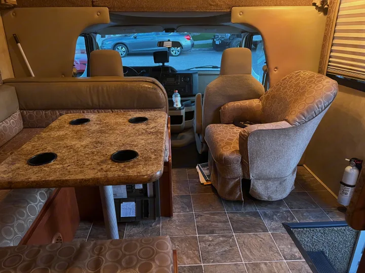 An RV rental interior looking forward, showing a dinette with cupholders, a comfortable recliner, and the cockpit with cars visible outside.. Thor Chateau 2012