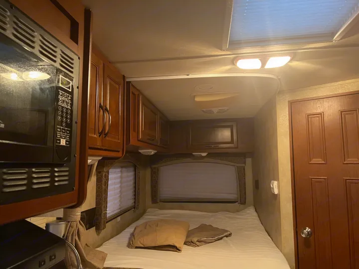 Inside a cozy RV rental: a bedroom and kitchenette area with a microwave, bed, wooden cabinets, windows, and ceiling lights.. Thor Chateau 2012