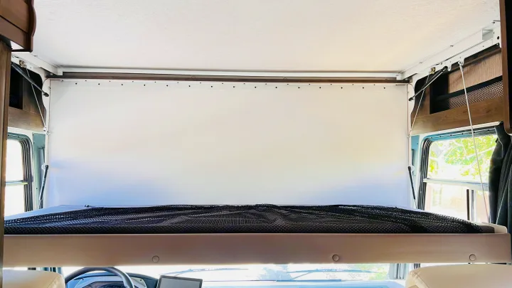An RV rental's front overhead bunk bed features a black safety net and white head panel, with the cockpit below.. Forest River Coachmen Pursuit 2022