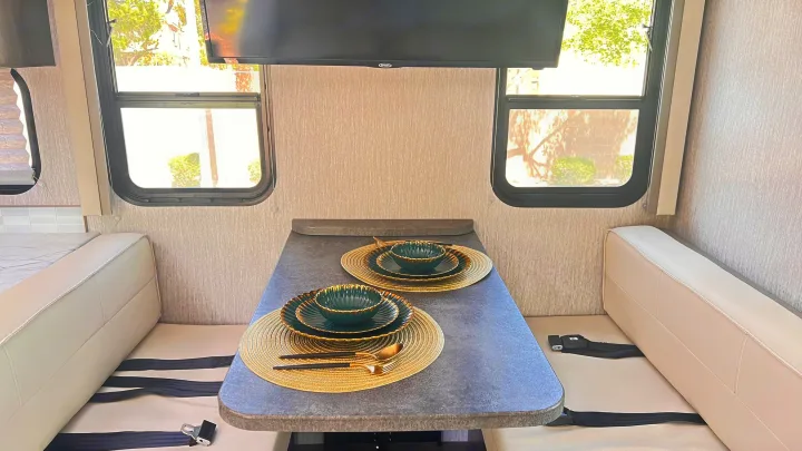 Cozy RV rental dining area showing a set table for two, comfortable bench seating with seatbelts, large windows, and an overhead TV.. Forest River Coachmen Pursuit 2022