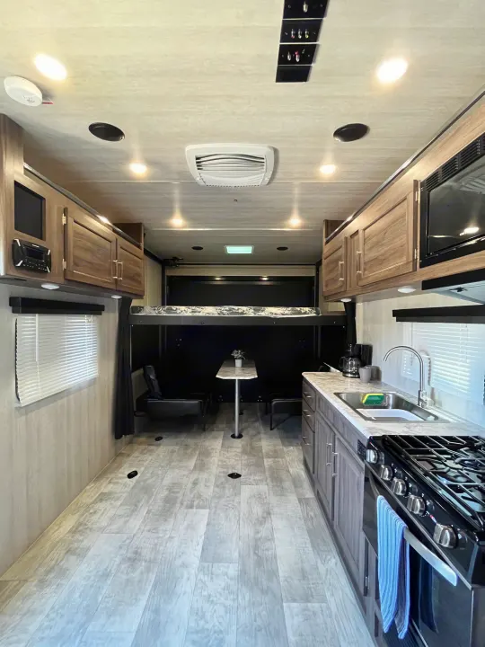 Bright RV rental interior: kitchen with stove/sink, dining table, and raised bunk bed above a versatile garage space.. Pacific Coachworks Blaze'N 2022