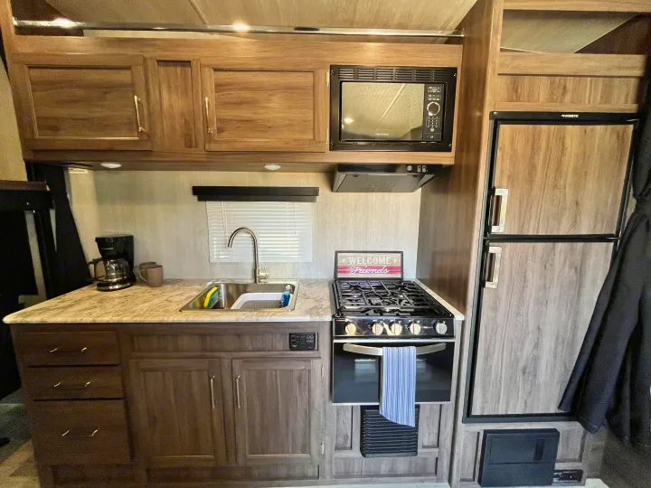 Inside an RV rental: a compact kitchen with sink, stove, oven, microwave, and Dometic refrigerator.. Pacific Coachworks Blaze'N 2022