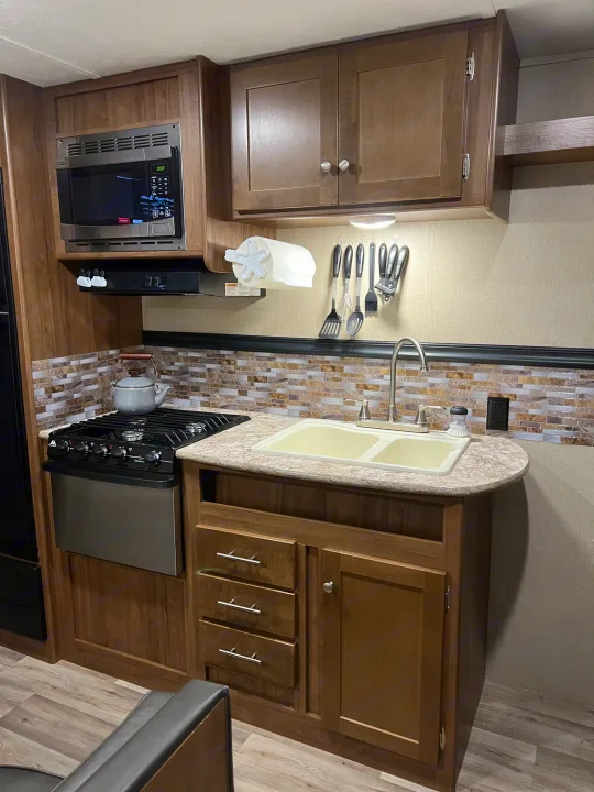 RV rental kitchen featuring a microwave, stove, double sink, wood cabinets, and tile backsplash.. Aspen Trail Aspen Trail Trailer 2018