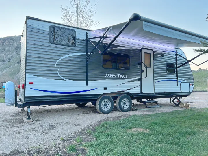 Aspen Trail RV rental travel trailer set up on gravel. Its awning is extended & illuminated, ready for camping.. Aspen Trail Aspen Trail Trailer 2018