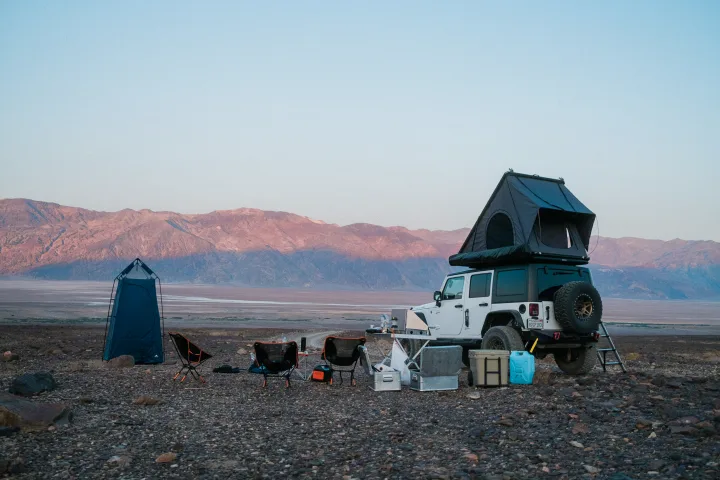 White Jeep with rooftop tent & camp setup (chairs, privacy tent) in a vast, rocky desert with mountains. Perfect for an RV rental trip.. Jeep Wrangler Rubicon 2017