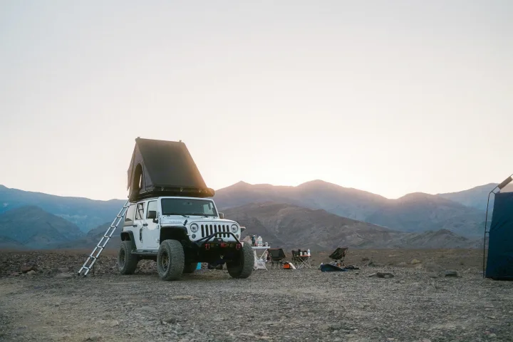 A white Jeep with a rooftop tent and camping gear in a vast, rocky desert with mountains, a unique RV rental alternative.. Jeep Wrangler Rubicon 2017