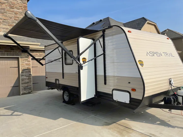 Sunny view of an Aspen Trail RV rental, showing its extended awning and open door, parked on a driveway.. Aspen Trail LE 2021