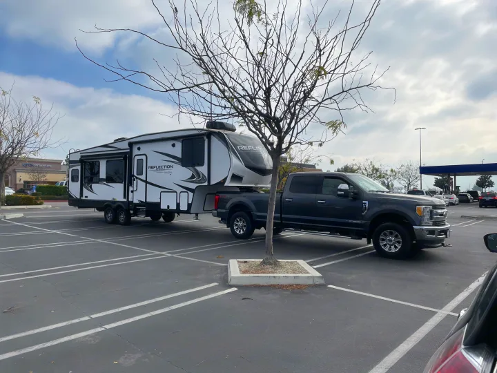 A dark grey truck tows a large white Reflection fifth-wheel RV in a parking lot, typical of an RV rental or personal rig.. Grand Design Reflection 2021