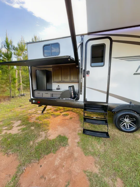 An rv rental showing its open outdoor kitchen with a fridge, sink, and grill. Steps and awning extended for camping enjoyment.. Cruiser RV MGP 3100BH 2022