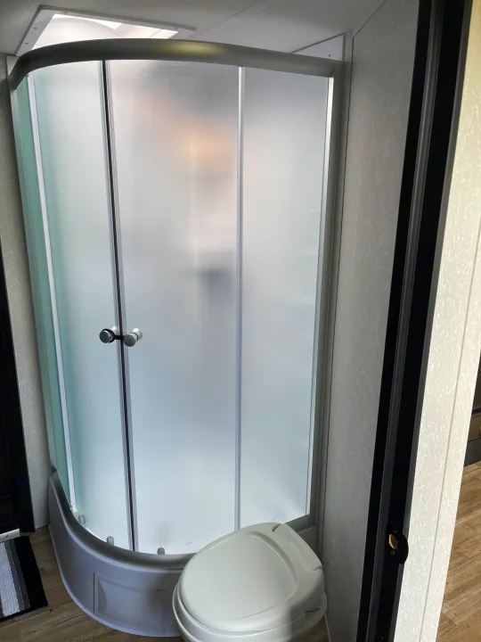 An RV rental bathroom with a rounded frosted glass shower enclosure and a white toilet on wood-look flooring.. Forest River Cherokee Grey Wolf 2022