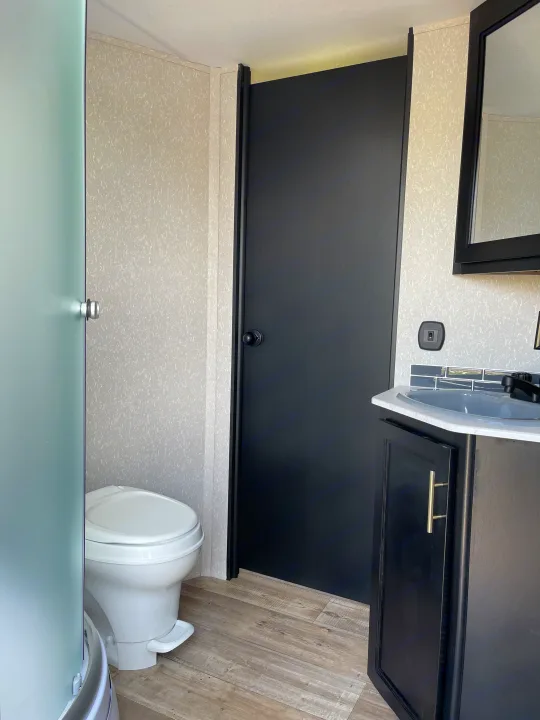 An RV rental bathroom with a white toilet, black sink vanity, and frosted shower door. A black door is on the right.. Forest River Cherokee Grey Wolf 2022