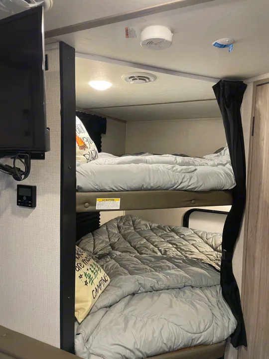 Cozy bunk beds in an RV rental interior, featuring light gray bedding, a mounted TV, and a privacy curtain. Ideal for camping.. Keystone RV Passport SL 2021