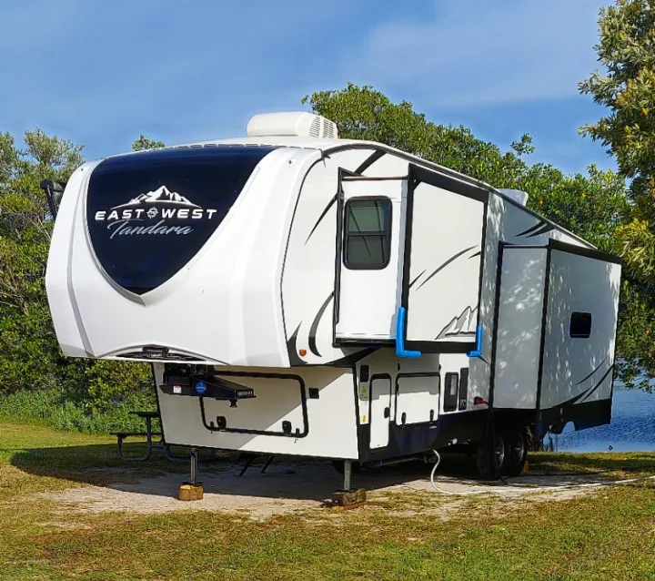 Large white East West Tandara fifth-wheel RV, parked on a grassy site by water, shows its extended slides. Consider an rv rental.. East to West Tandara 2021