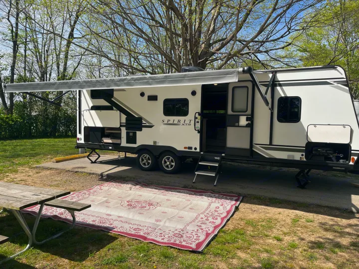 An RV rental set up at a sunny campsite with its awning extended, an outdoor rug, and a picnic table. Perfect for nature lovers.. Coachmen 2549 BHX 2021