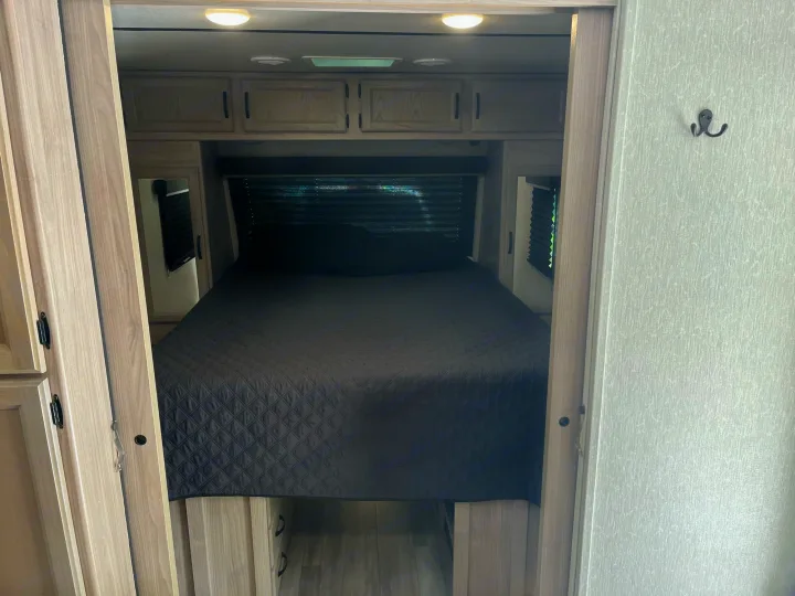An RV rental bedroom interior, featuring a dark grey bed, overhead cabinets, mirrored side closets, and window blinds.. Coachmen 2549 BHX 2021