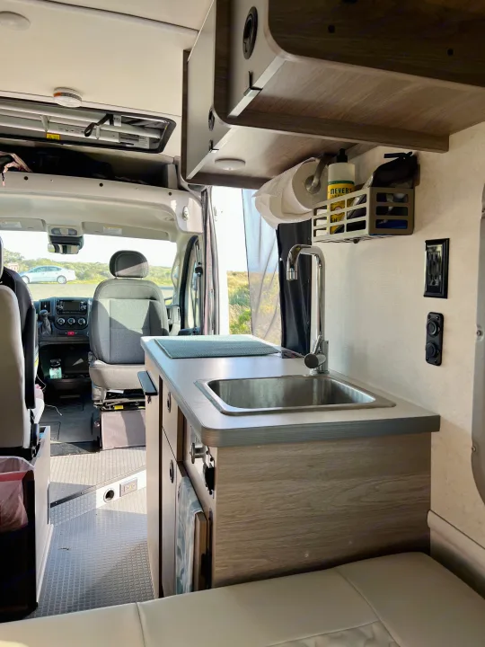 RV rental interior: compact kitchen with sink, paper towels, cleaning supplies, driver's cab, and seating.. Winnebago Solis BUT59P 2024