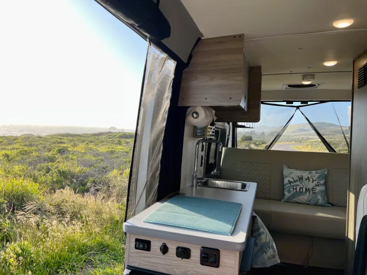 RV rental interior: A compact kitchen and seating area with a screen door open to a sunny coastal view with green grass and ocean.. Winnebago Solis BUT59P 2024
