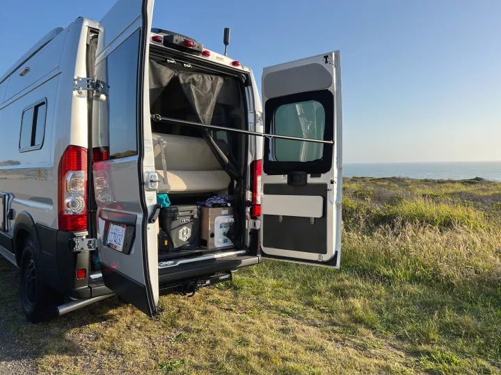 Silver RV rental camper van parked on a grassy cliff, back doors open revealing interior and ocean view. Enjoy scenic travels!. Winnebago Solis BUT59P 2024