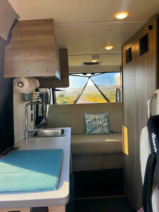 Cozy RV rental interior with kitchen sink, seating area, and a pillow reading "ALWAYS HOME." Scenic view through the rear screen.. Winnebago Solis BUT59P 2024