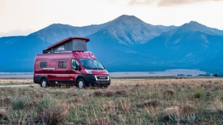 Explore nature! This red RV rental camper van, with its pop-up top, is parked in a scenic field before blue mountains.. Winnebago Solis 2021