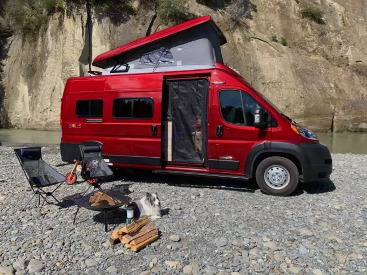 Experience van life! Red pop-top rv rental parked by a river, with chairs, fire pit, and a dog for ultimate outdoor adventure.. Winnebago Solis 2021