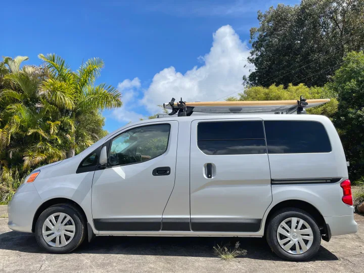 Compact RV rental: A white van with a surfboard on its roof, parked under a blue sky with vibrant palm trees, ready for adventure.. Nissan NV200 2021