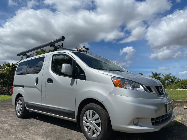 Compact silver Nissan NV200 RV rental camper van with rooftop awning and watersports gear under a sunny sky.. Nissan NV200 2021