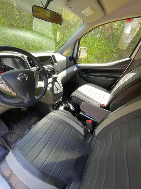 Interior of a Nissan van, perfect for your next RV rental adventure. Shows driver's cockpit, passenger seat, and green foliage outside.. Nissan NV200 2021