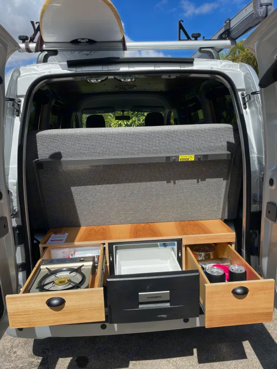 This RV rental features a compact pull-out kitchen with stove, fridge, and a surfboard on the roof rack. Adventure-ready!. Nissan NV200 2021