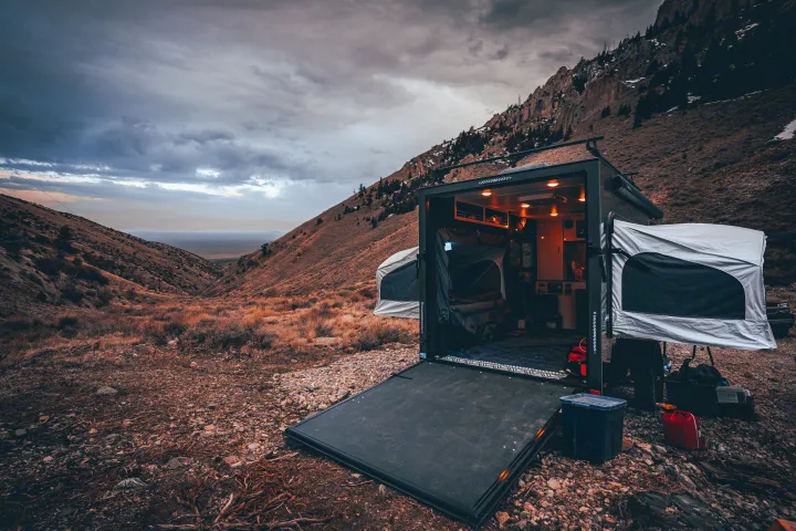A custom off-road trailer, resembling an RV rental, open in a rugged mountain valley, revealing a cozy, lit interior.. InTech RV Flyer Discover 2021