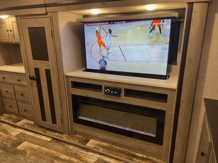 Cozy RV rental entertainment setup: A large TV shows basketball above an electric fireplace, with sleek cabinets & drawers.. Keystone Outback 2020