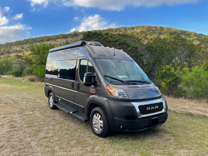 This grey Ram ProMaster RV rental is ready for adventure, parked on grass before scenic hills & sky.. Thor Scope 18M 2022