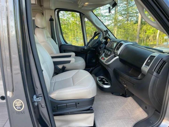 Inside an rv rental: inviting front cabin with comfortable beige seats, modern dashboard, and a view of green trees.. Thor Scope 18M 2022