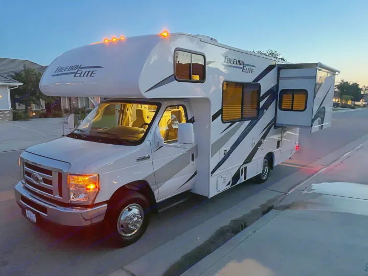 White Freedom Elite RV rental parked on a street at dusk, with lights on and a slide-out extended.. THOR MOTOR COACH FREEDOM ELITE 22fef 2022