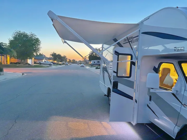 White RV rental parked on a quiet street at sunset, with its awning extended and interior lights glowing.. THOR MOTOR COACH FREEDOM ELITE 22fef 2022