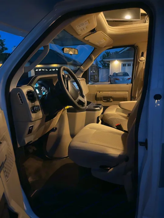 Inside an RV rental van: The well-lit driver's cabin with steering wheel, dashboard, and passenger seats, parked at dusk.. THOR MOTOR COACH FREEDOM ELITE 22fef 2022