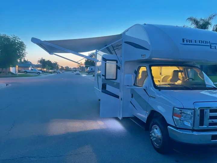 A Class C rv rental sits on a street at dusk, its awning extended and interior lights glowing, inviting adventure.. THOR MOTOR COACH FREEDOM ELITE 22fef 2022